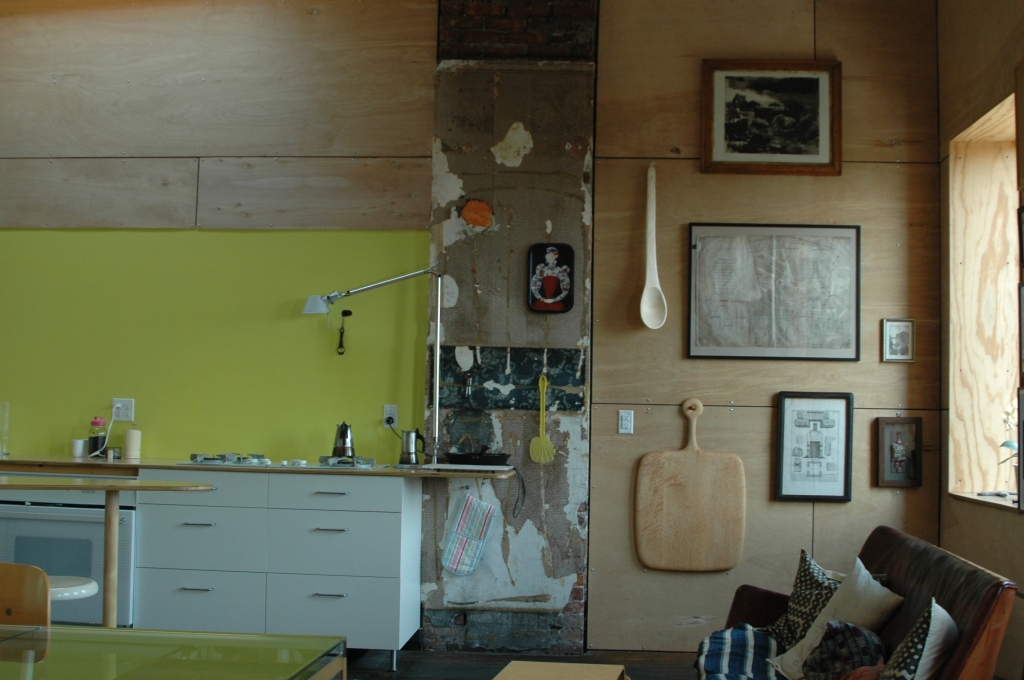 an aimge of Arscott's kitchen - there are bare, wooden walls where photos, a cutting board, and a wooden spoon hang. The backsplash for the counter area is a bright neon yellow