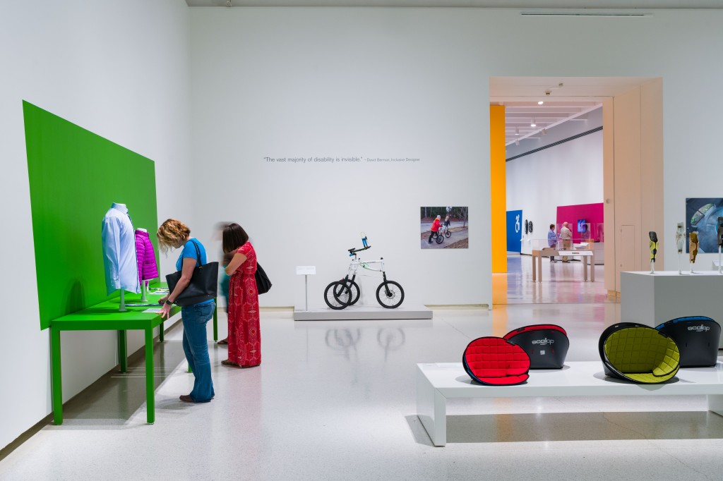 visitors stand in a white gallery, where a bright green painted wall highlights accessible clothing. the gallery also has seating and biking examples on white platforms