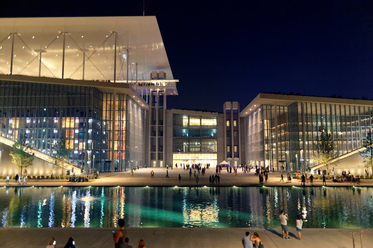 Stavros Niarchos Foundation Cultural Center - a large structure with glass facades with a shallow reflecting pool out front. Warm light pools out from the buildings, and people gather and wander outside