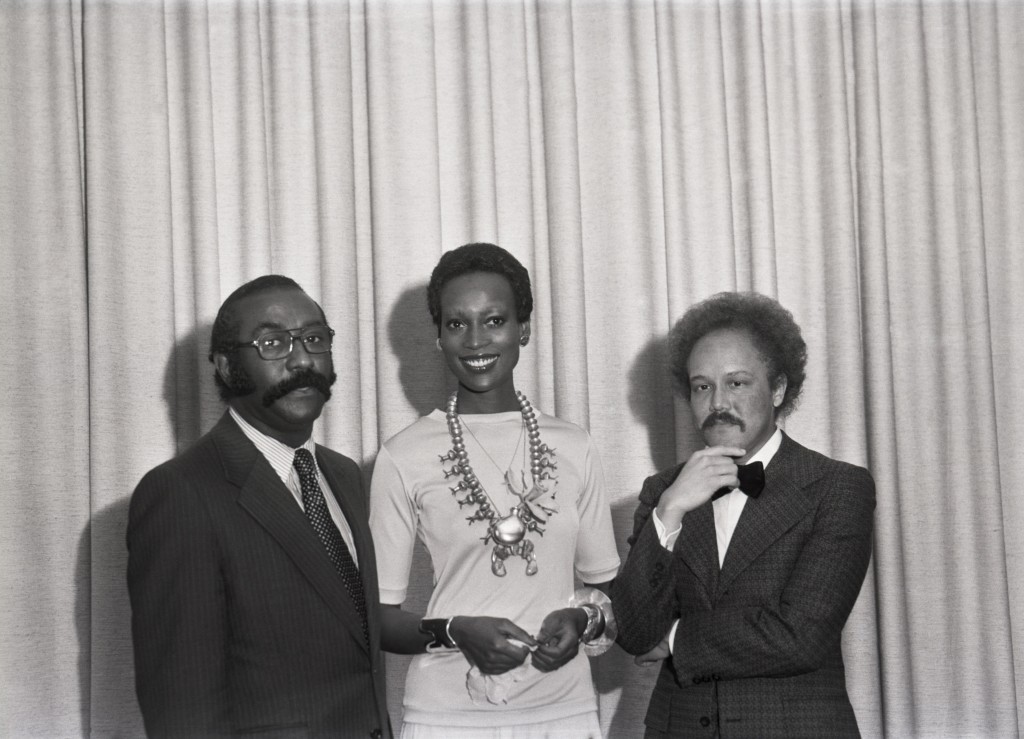  a black and white photo of Naomi sims, a black woman with a small fro, elegantly standing in between De, reed and Morris - both in suits